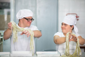 Male and female coworkers in 20s and 40s with Down Syndrome smiling at one another as they lift freshly made fettuccine into boxes.